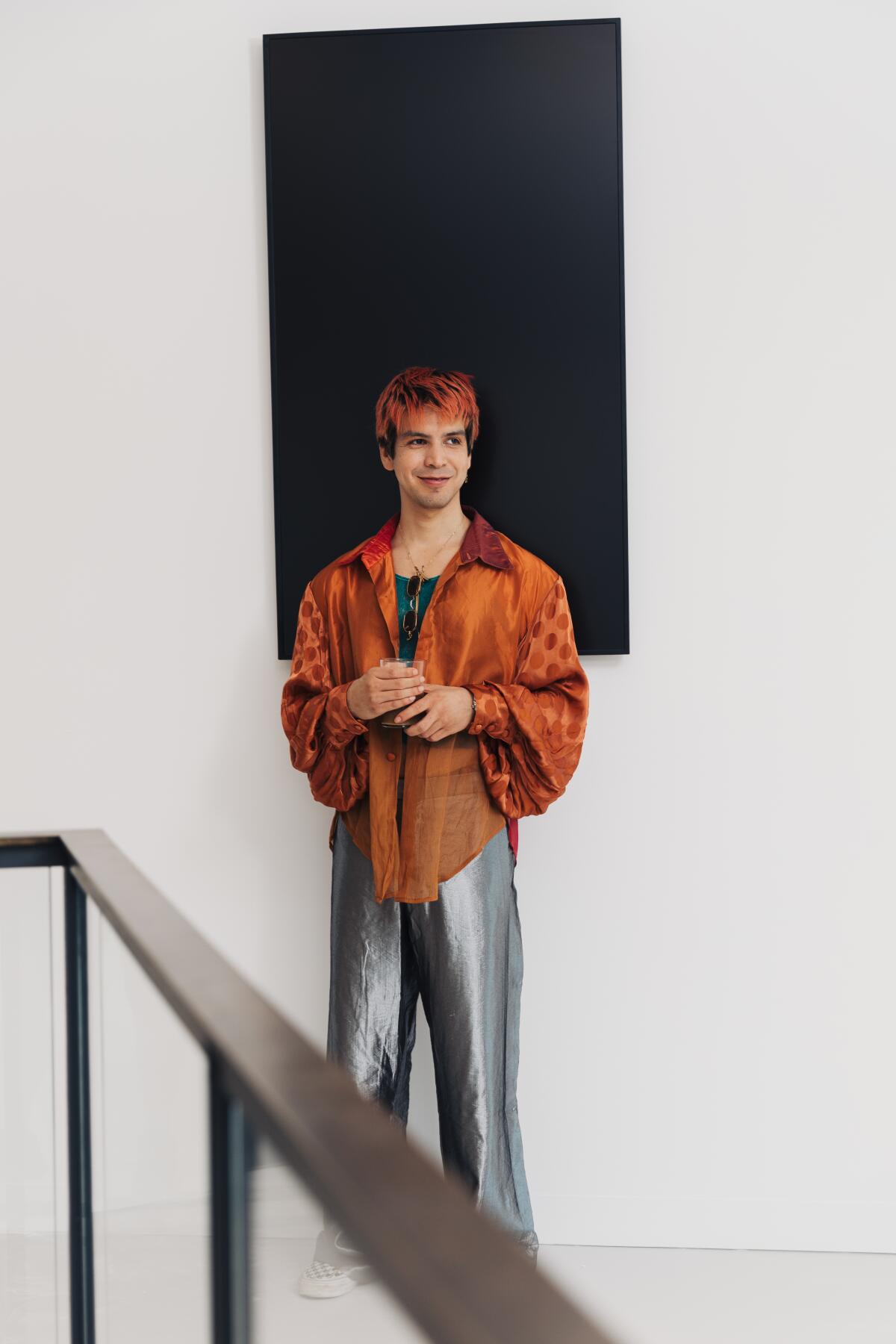 Julio Torres, in silky gray pants and an orange shirt, stands before a black painting in a white room.