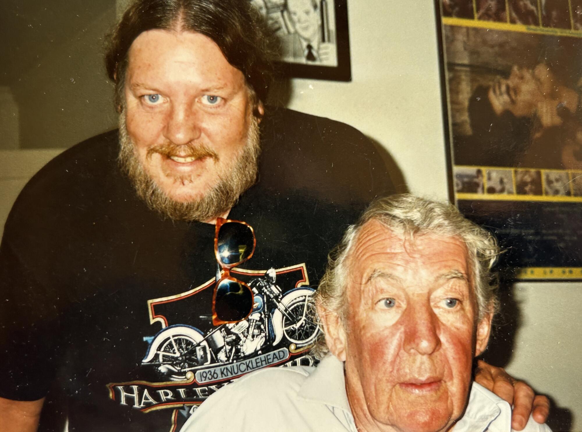 A seated older man with a bearded man standing behind him