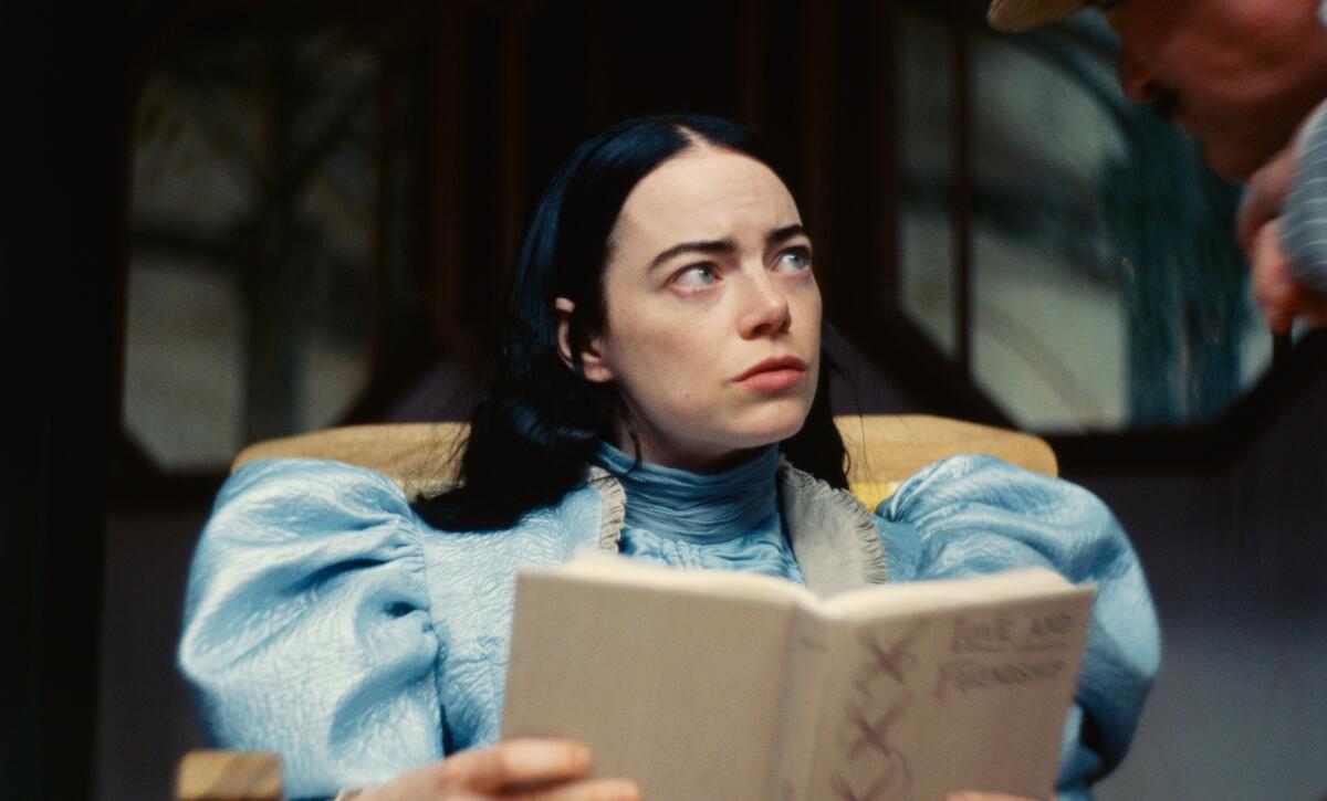 Emma Stone wears a light blue dress with large puff sleeves while reading a book in a still from "Poor Things."