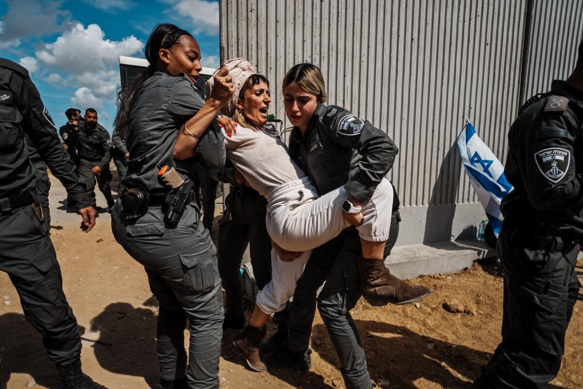 Two soldiers picking up a struggling woman near a tall security wall as other troops stand by