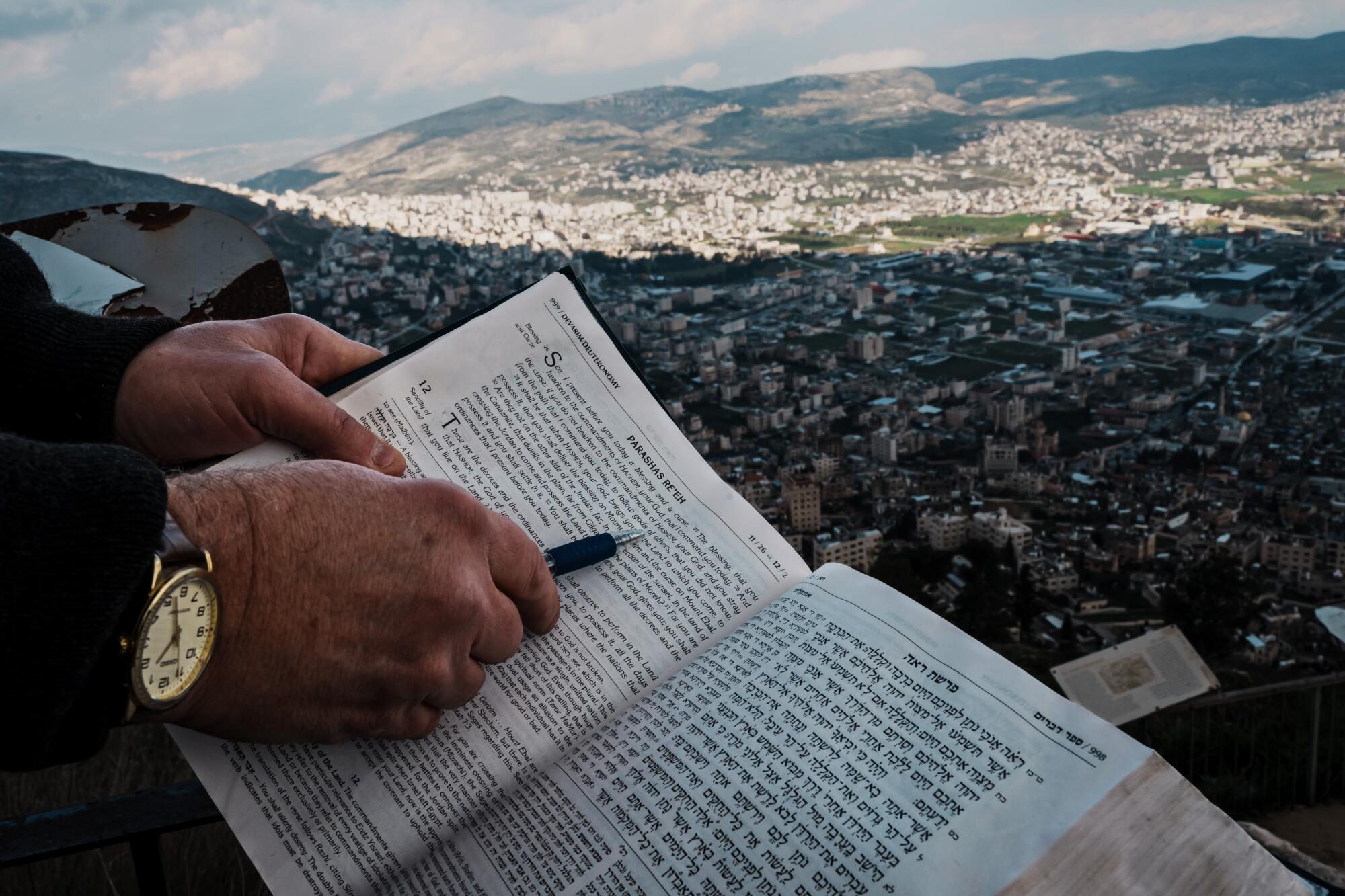 A hand pointing to a passage in English in a Hebrew Bible, a sprawling community in the valley below 