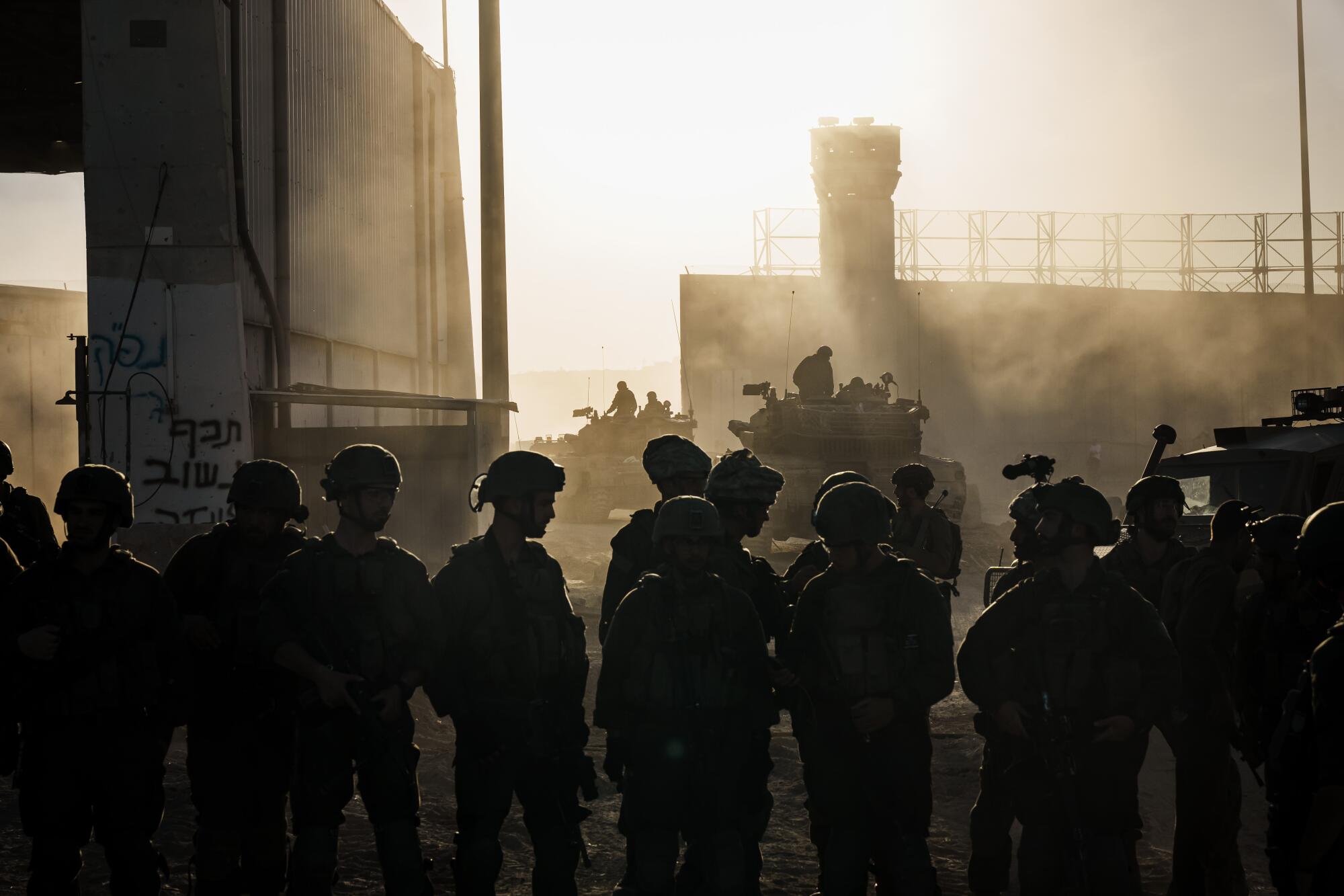 About a dozen soldiers standing in the haze as others operate tanks near a security wall