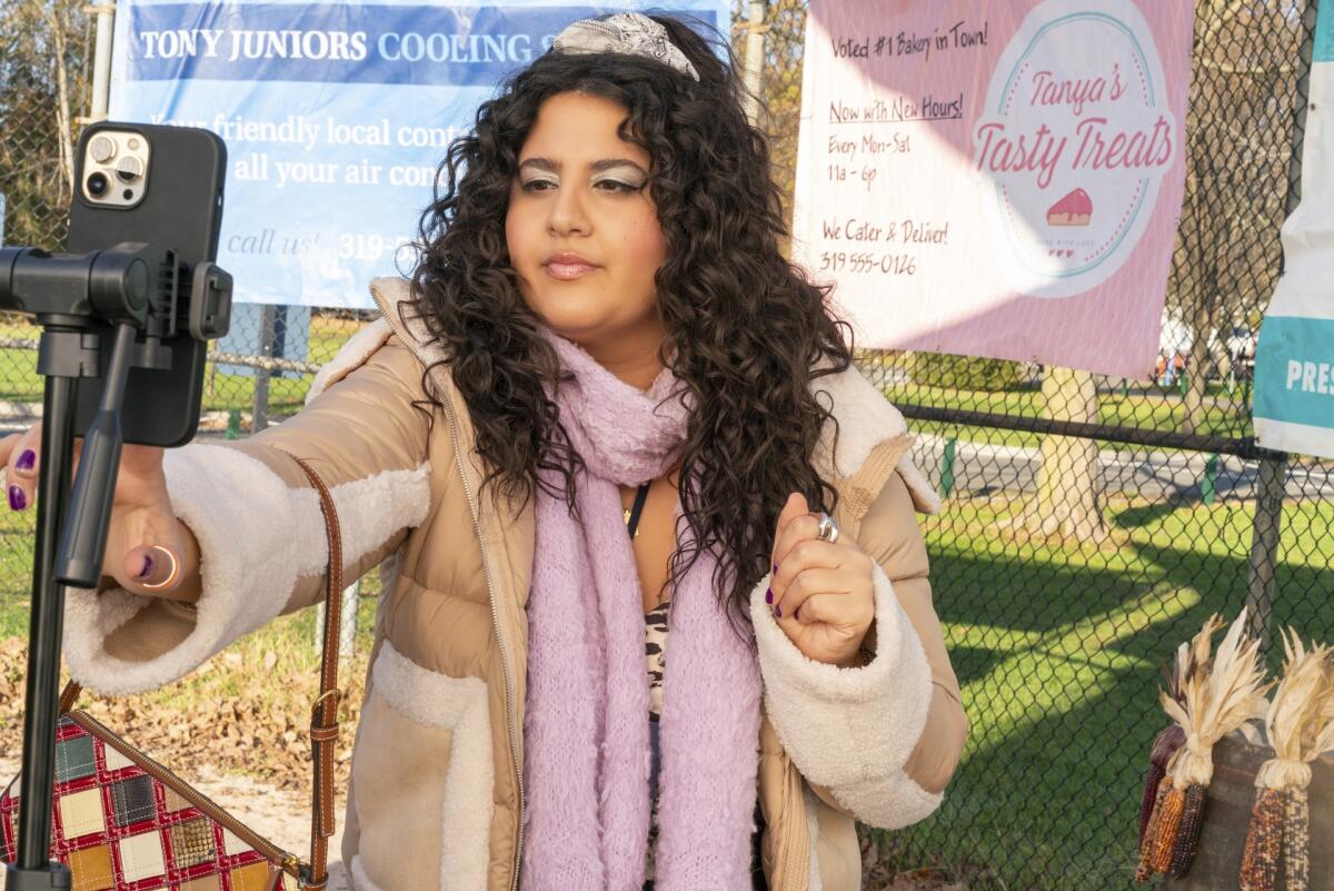 A woman in a coat and pink scarf looks toward a phone on a tripod.