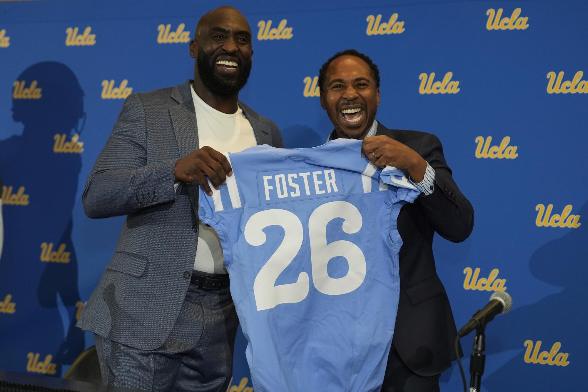 New UCLA head football coach DeShaun Foster holds up a Bruins jersey while posing with athletic director Martin Jarmond 