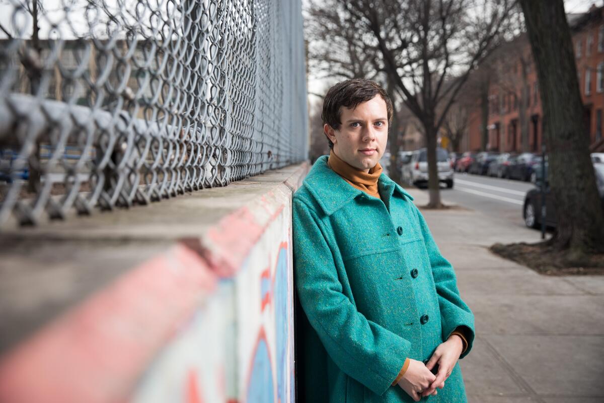 Cole Escola leans against a concrete and chain-link fenced wall.