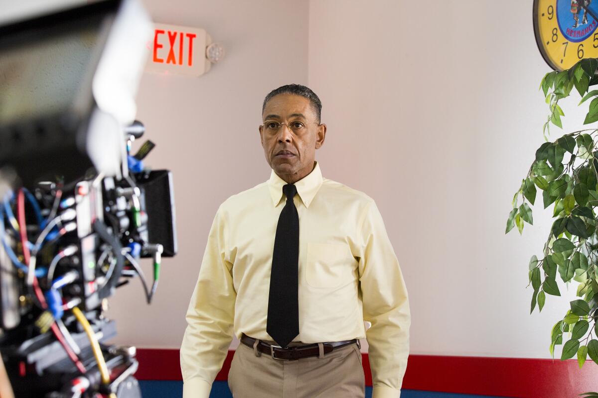 A man wearing a yellow dress shirt and black tie stands in front of a camera.