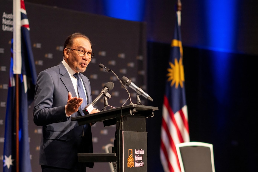 An elderly Asian man in a suit gestures as he gives a talk at a rostrum with flags behind