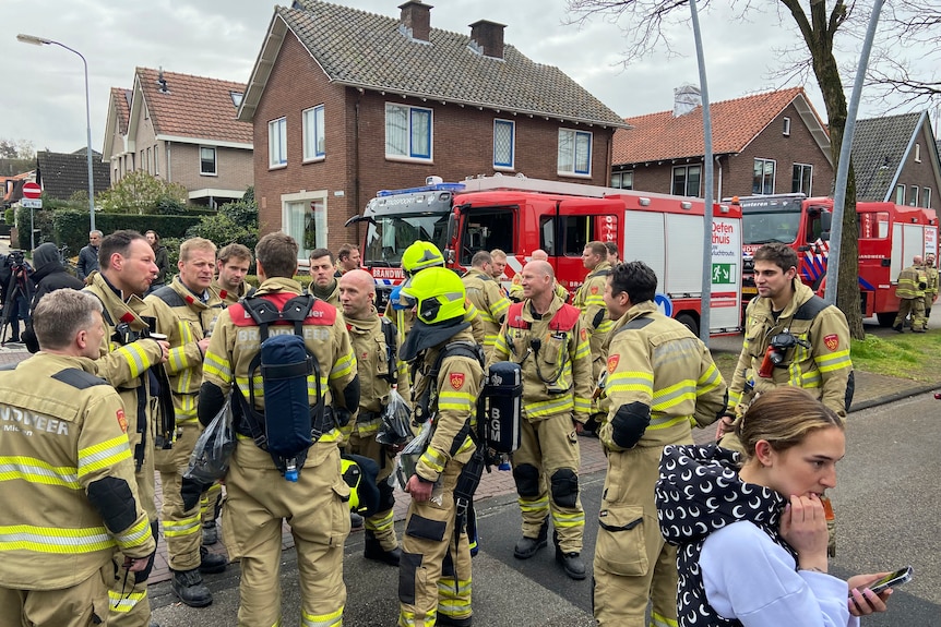 A gathering of firefighters in a small town with red brick houses in the background