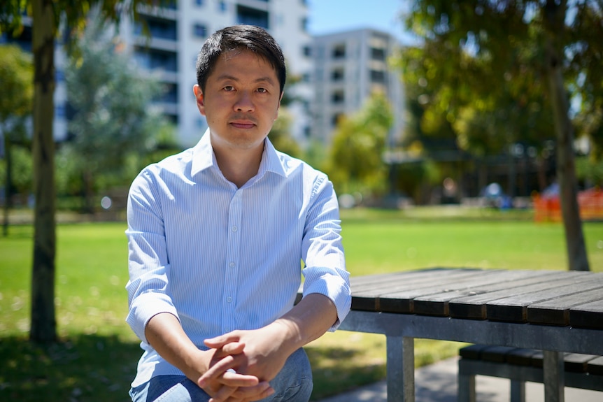 A man in a business shirt sits on a park bench surrounded by trees 