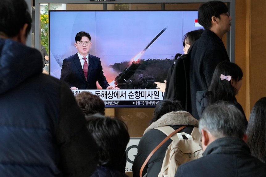 A TV shows and Asian newsreader reporting a rocket launch as people sit in a room and watch