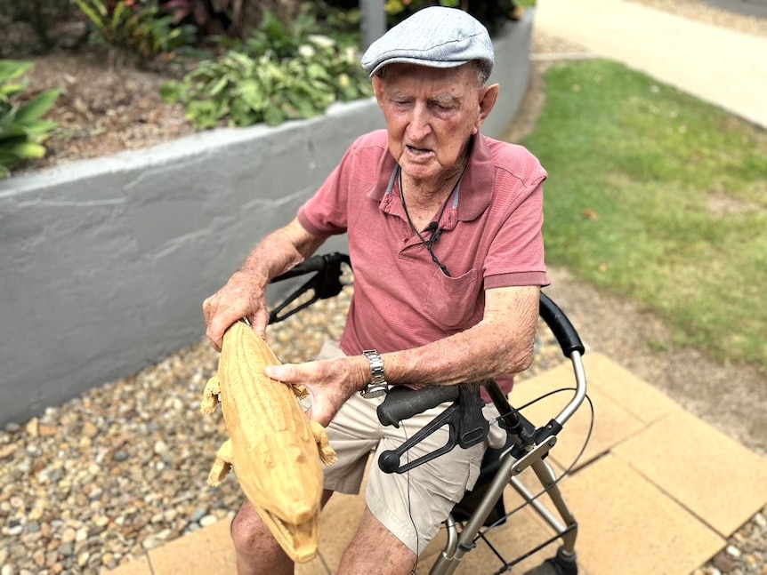 An older man sitting on a walker holds up a wooden matchstick model of a crocodile.