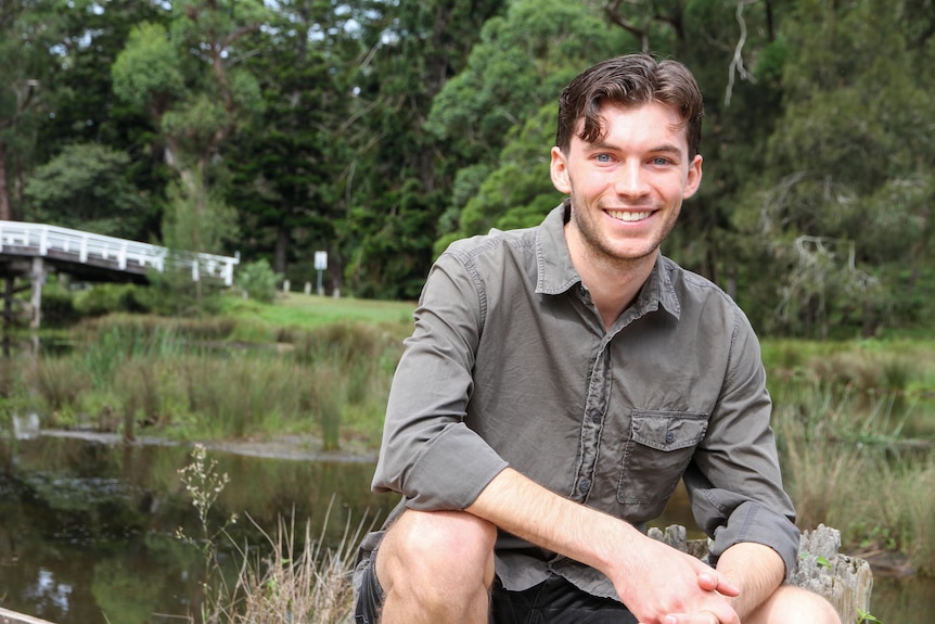 Man in his 20's with dark facial hair, wearing grey shirt, squatting in front of river