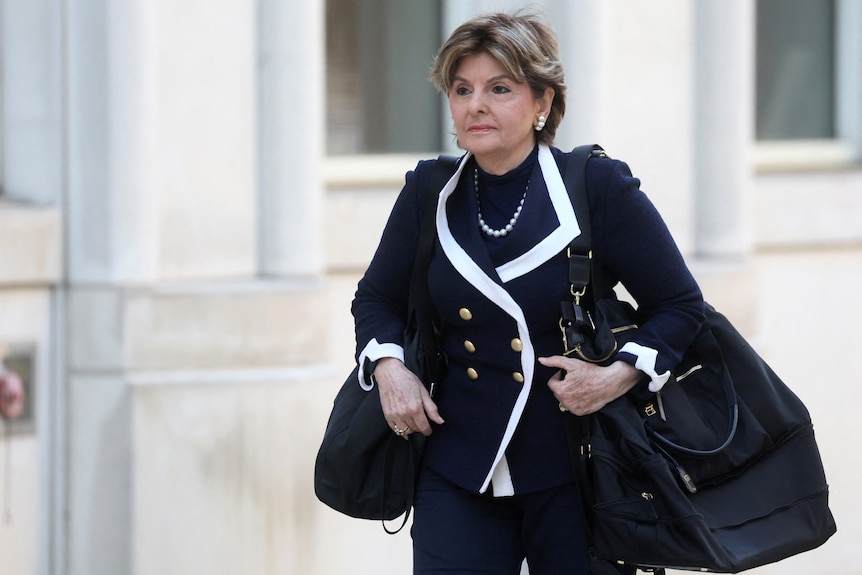 Gloria Allred walks outside wearing black and white and carrying larged bags.