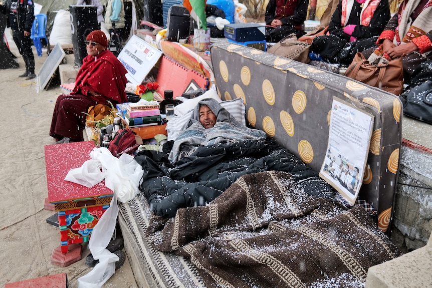 A man lays under grey blankets covered with light snow on a mattress outside 