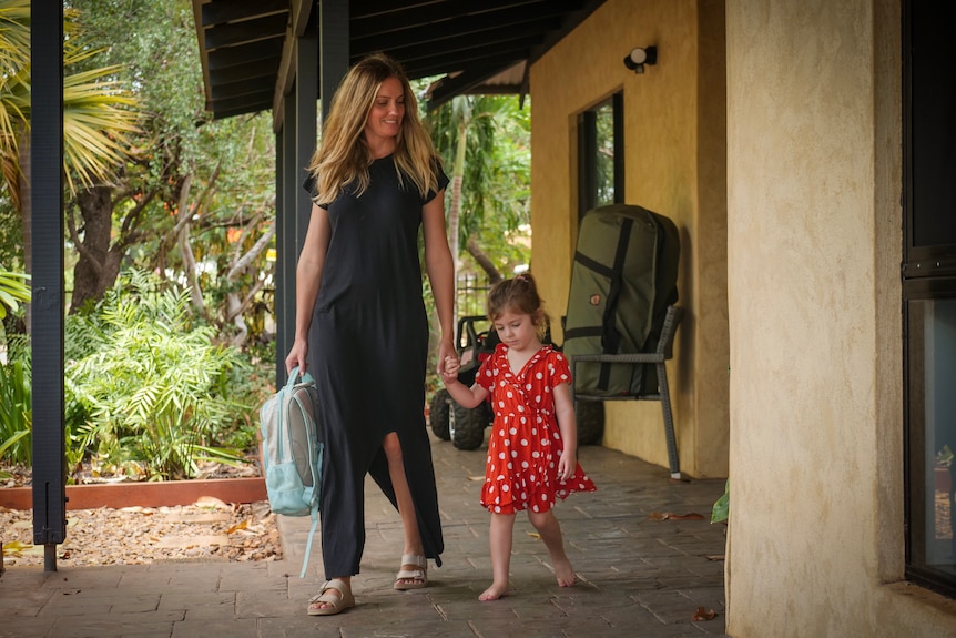 A woman holding her young daughter's hand and a blue backpack walking outside a home.