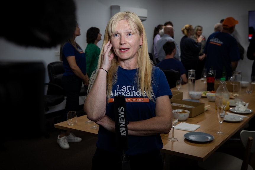 A woman with long blonde hair and a blue t-shirt holds a hand to her ear as she speaks to camera.