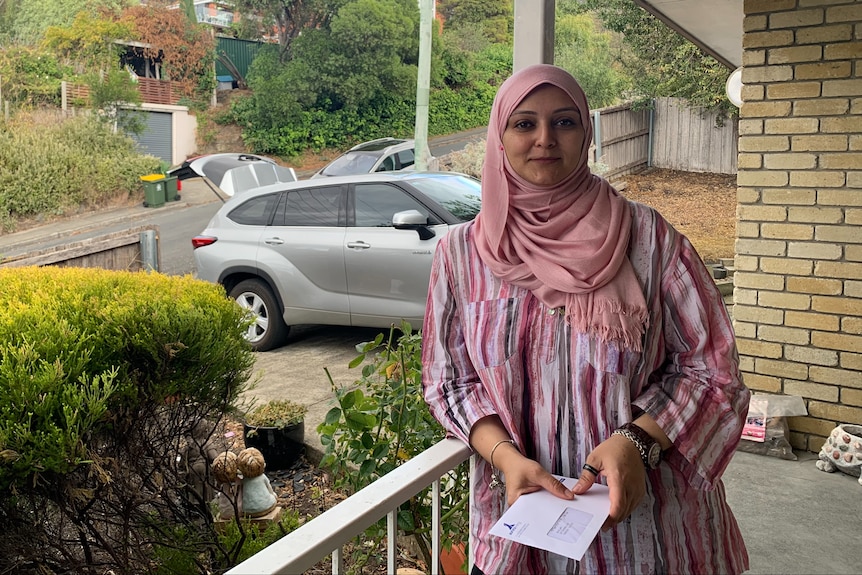 A woman in a pink headscarf smilse at the camera while standing on her porch