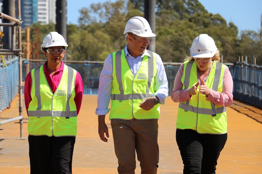 Three people in officewear, high vis and high hats walk and talk in daylight.