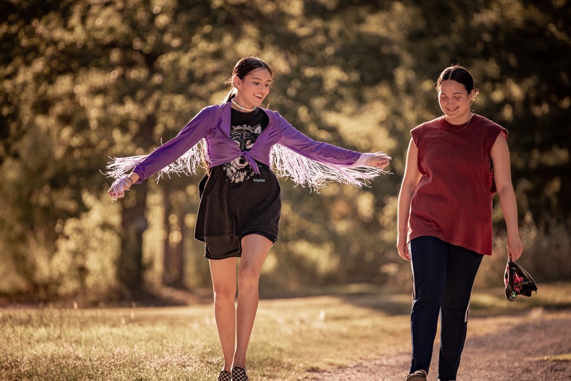 Lily Gladstone walking next to a girl who is wearing purple with feathers, they are outside and look happy