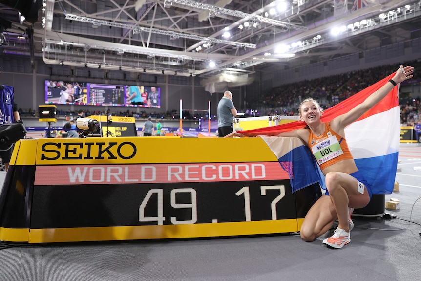 A Dutch athlete smiles and holds a national flag behind her back as she kneels next to a scoreboard with 'World Record 49.17'.