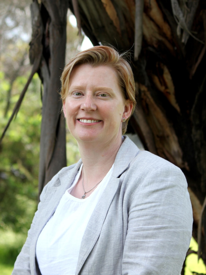 A woman with short red hair standing in front of a tree trunk. 