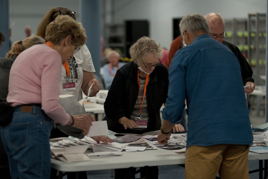 A group of five people stand over a table with stern faces as they count votes. 