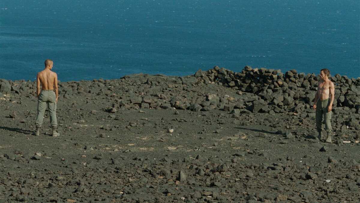 Two shirtless men stand on a craggy shore.