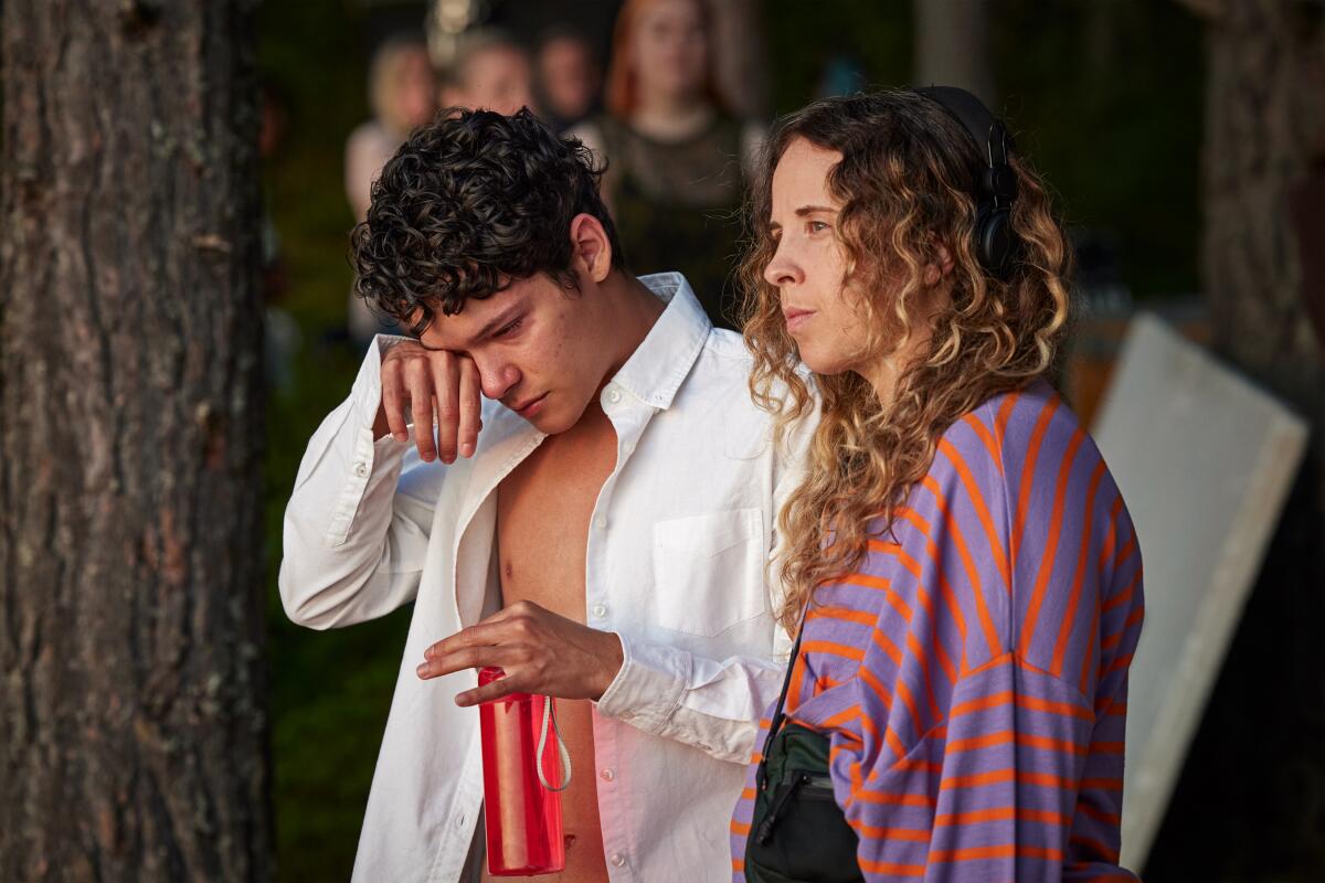 A boy wearing an open white shirt rubs the corner of his face as a woman stands next to him.