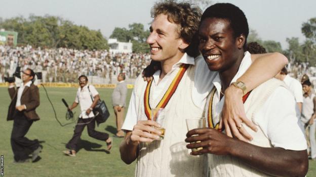 Jonathan Agnew and Norman Cowans walk off the pitch holding glasses of champagne