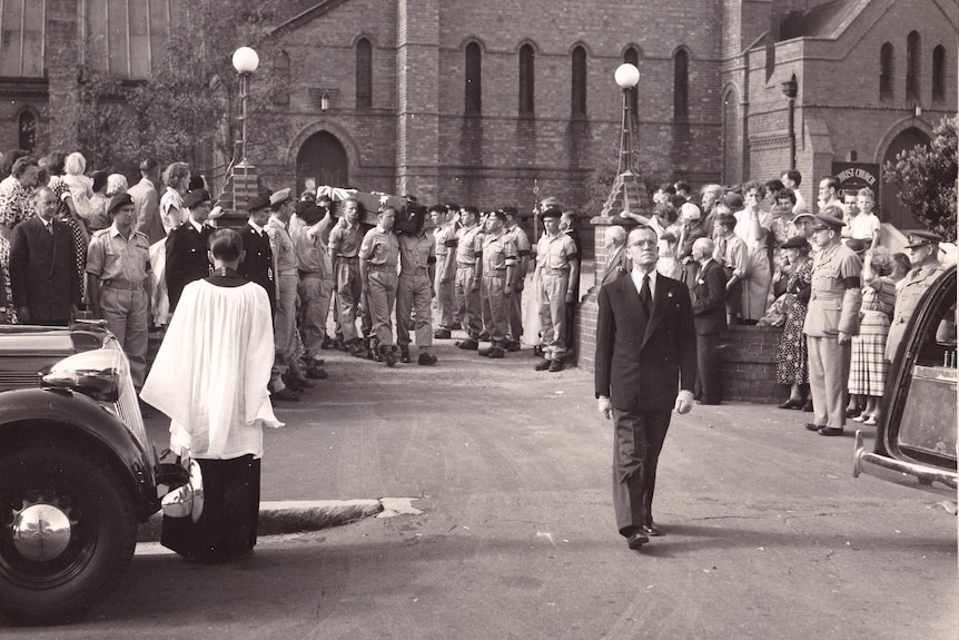 An old photo of a crowd watching on as a coffin is escorted from a church.