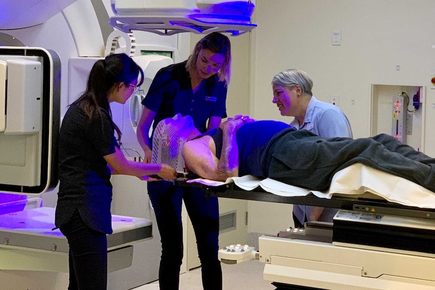 Man lies prone on table of hospital machine with two staff helping him