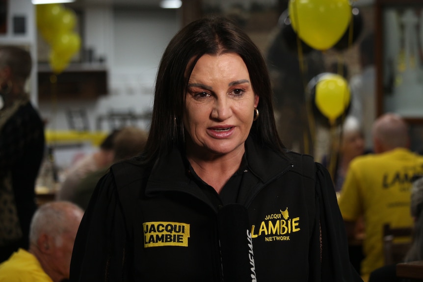 A woman with long, dark brown hair speaks to the camera with a serious expression. 