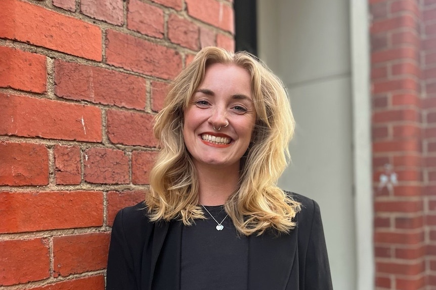 A blonde white woman wearing a black shirt and jacket. She's standing and smiling in front of a brick wall