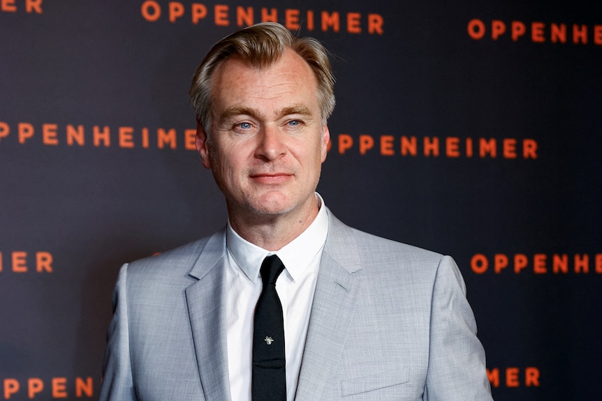 A man wearing a grey suit poses for a photo in front of a background that says Oppenheimer on it.