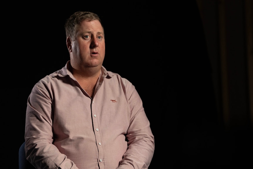 A man in a pink collared shirt sits in a dark room during an interview.