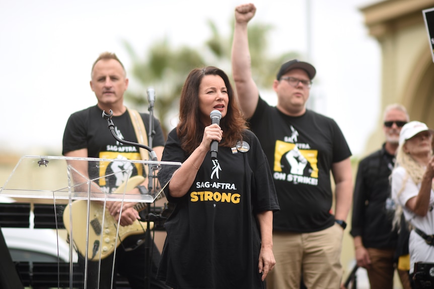 A woman standing and speaking into a microphone on stage at a rally. A man with a guitar and a man raising his fist behind her