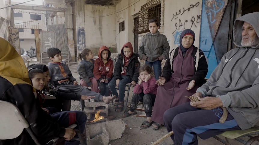 A woman and a man children sit gathered around a fire with their children in the rubble of a damaged home.