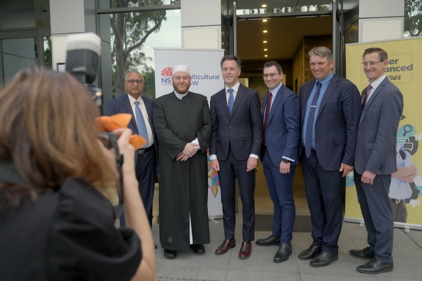 Six men in suits stand in front of photographer smiling