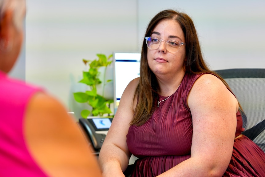 A white woman with brown hair and glasses listening to a woman speak. 