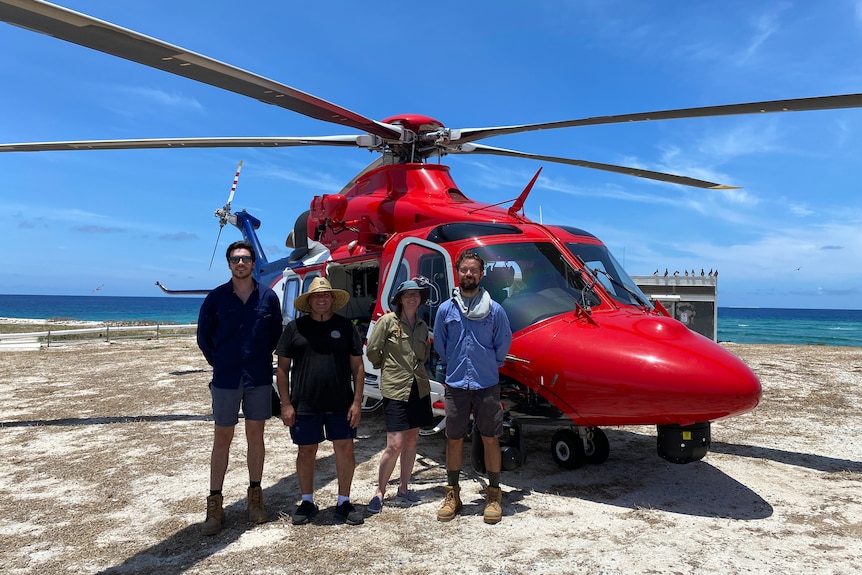 A woman and three men stand in front of a red helicopter near the ocean