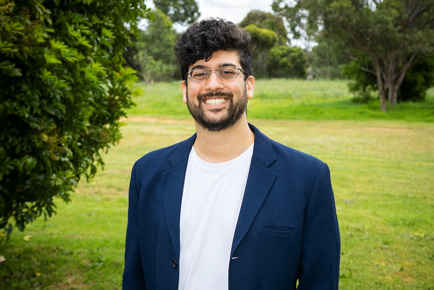 A man wearing glasses and smiling to camera with trees and grass in the background