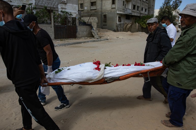 people carry a body wrapped in a white sheet with red flowers on it