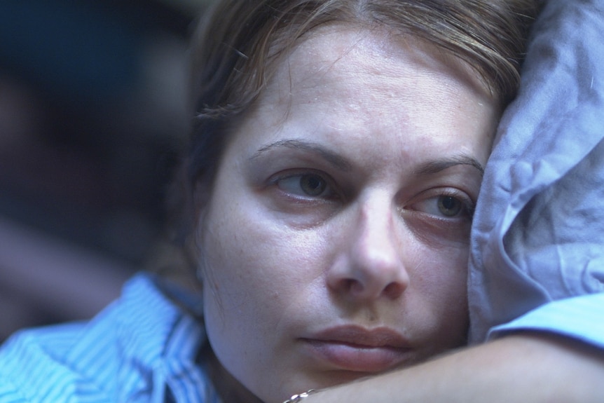 A woman rest her head on a pillow, she looks to the side, with a neutral expression.