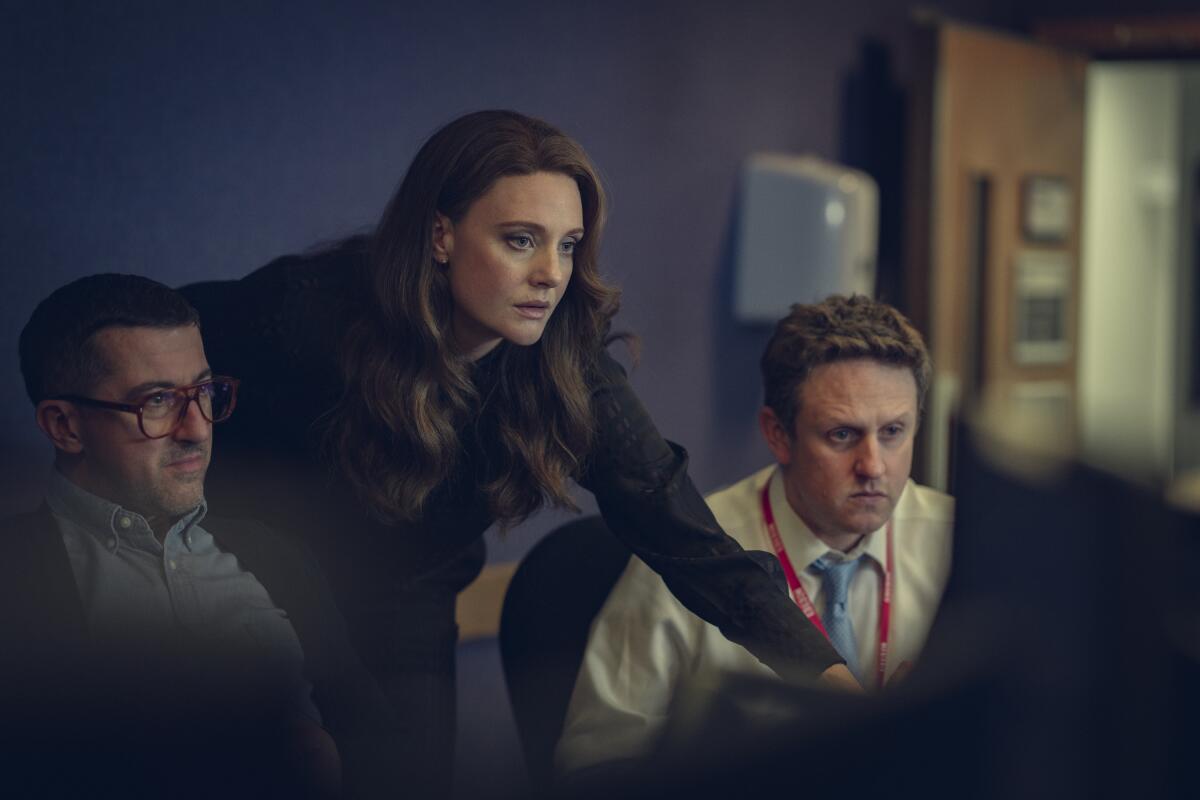 A woman, flanked by two men, leans to look at a screen on a desk.