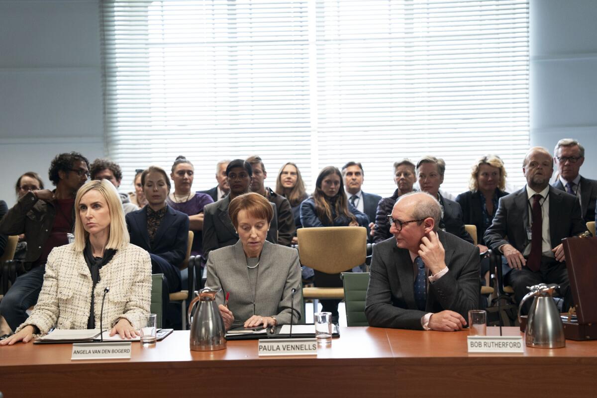 Three people sit a long table with microphones and documents in front of them.