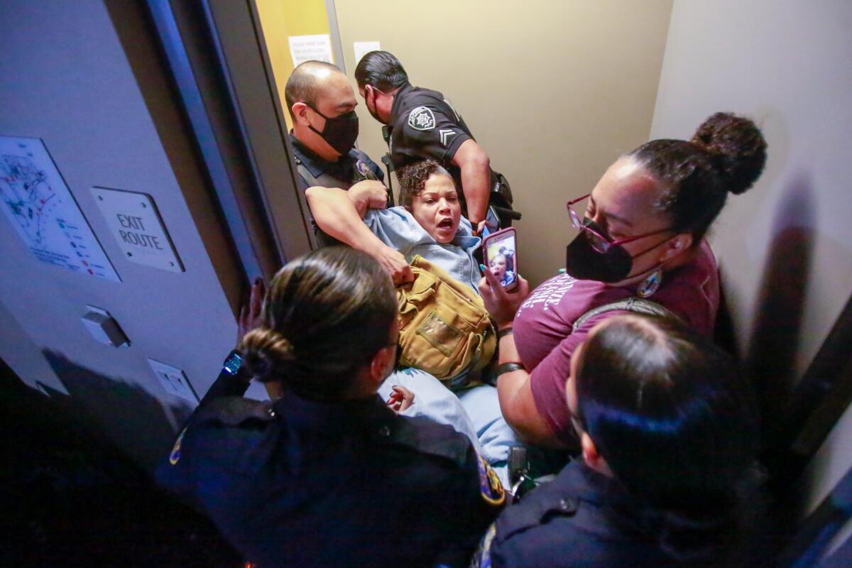 Two police officers carrying Melina Abdullah out of a room as others stand by