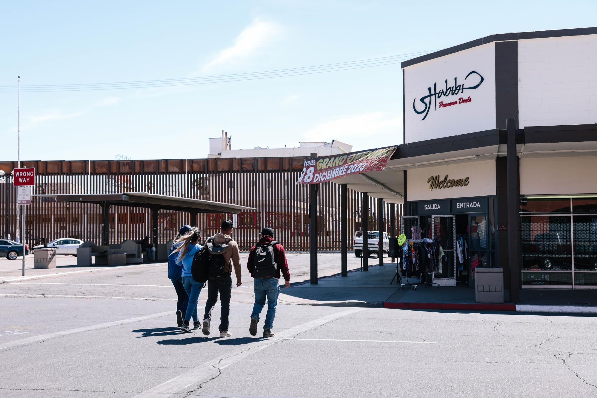 A group of people cross the street near a U.S.-Mexico border fence. 