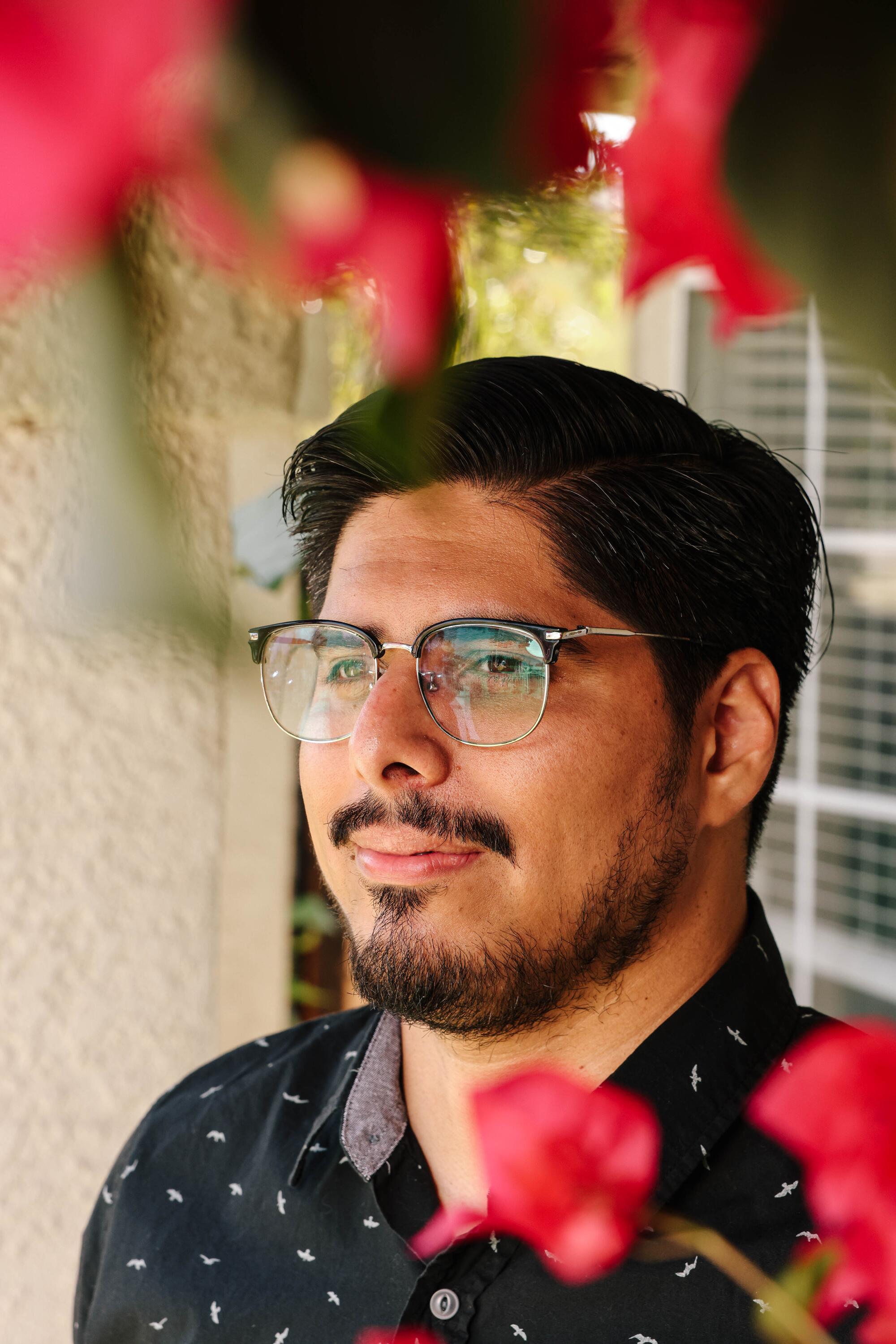 A bearded man in glasses and a black shirt poses amid red blossoms. 