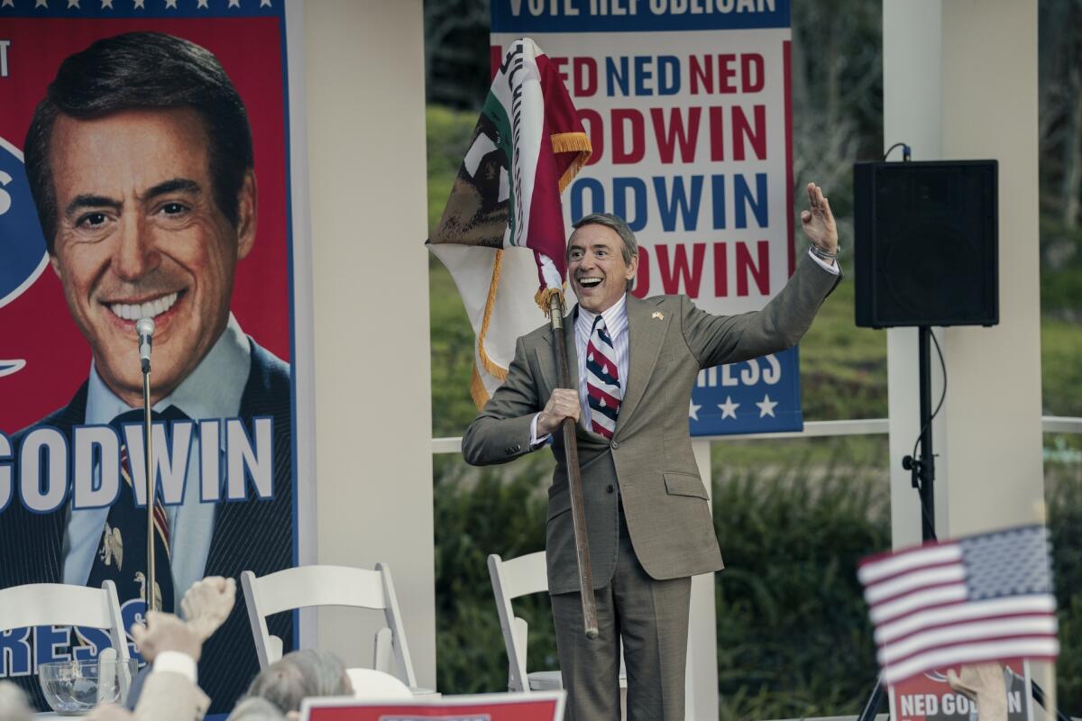 A man in a suit stands on a stage with signs behind him.