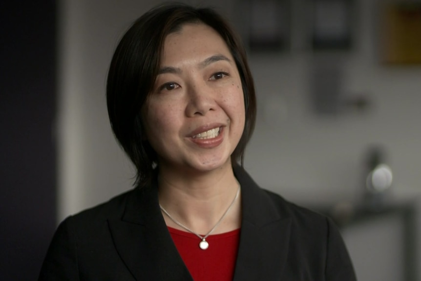 Woman with short hair wearing a red shirt and black jacket, sitting in an office.